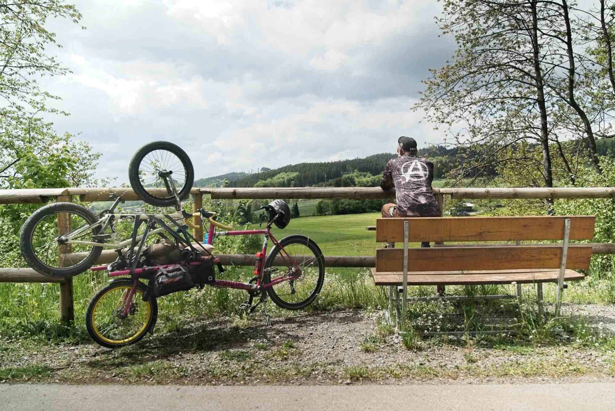 Bike-bikepacking: drei fahrräder, zwei leute und der sauerlandradring 10 Christian sitzt auf einer bank an einem schönen aussichtspunkt, das rad nebenan abgestellt